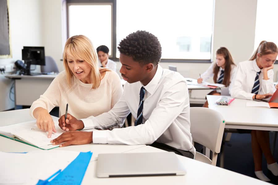 Boy sits in a classroom with his teacher helping him work.